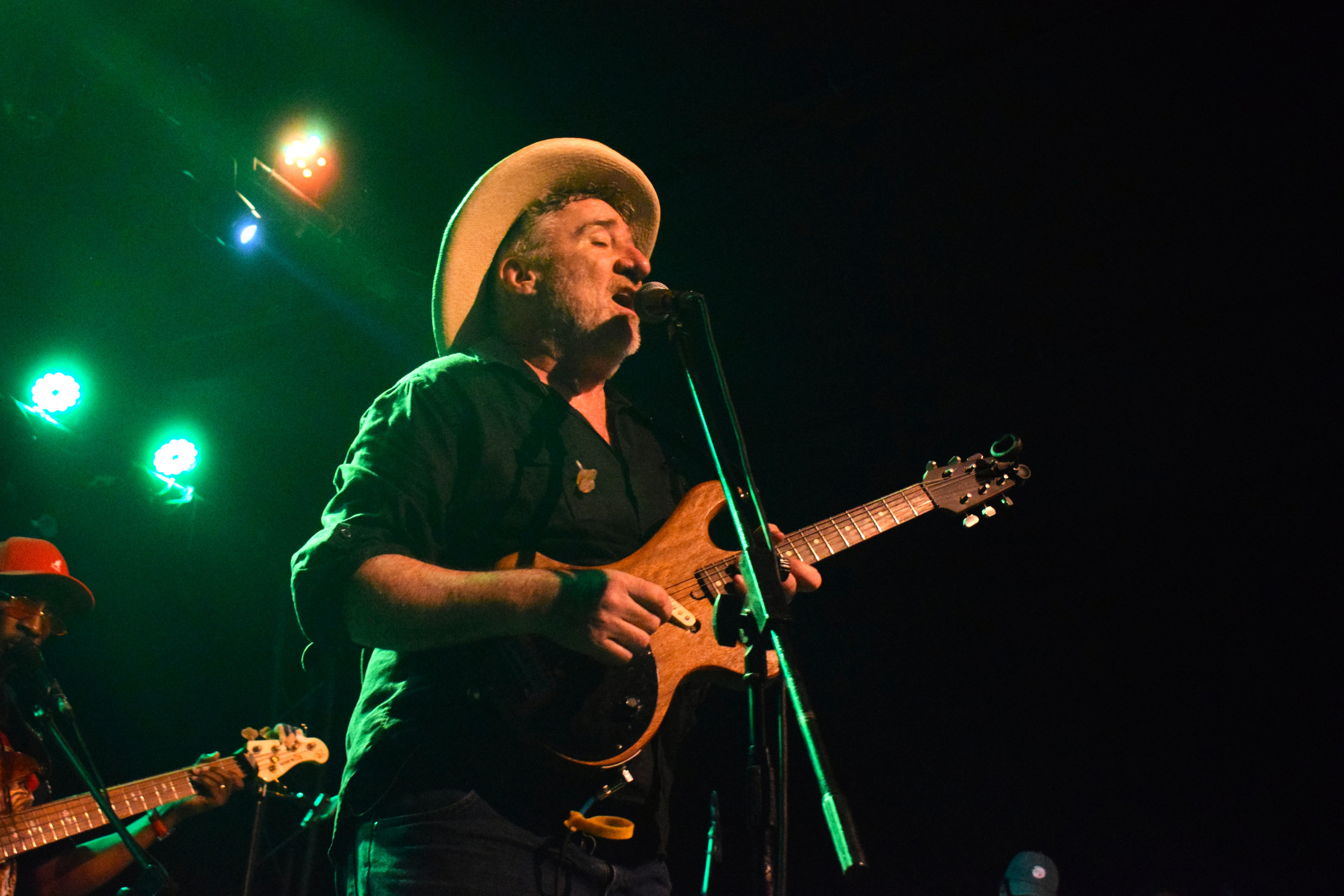 A male musician singing into a microphone and playing an electric guitar under stage lights, wearing a dark shirt and a light-colored cowboy hat.