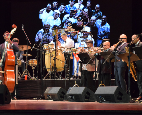 A large orchestra or big band performance on a stage, featuring musicians playing a double bass, drums, congas, trumpet, saxophone, and other brass instruments. A large projected image of smiling musicians is visible on the backdrop. A Cuban flag is visible behind the percussion.