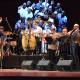 A large orchestra or big band performance on a stage, featuring musicians playing a double bass, drums, congas, trumpet, saxophone, and other brass instruments. A large projected image of smiling musicians is visible on the backdrop. A Cuban flag is visible behind the percussion.