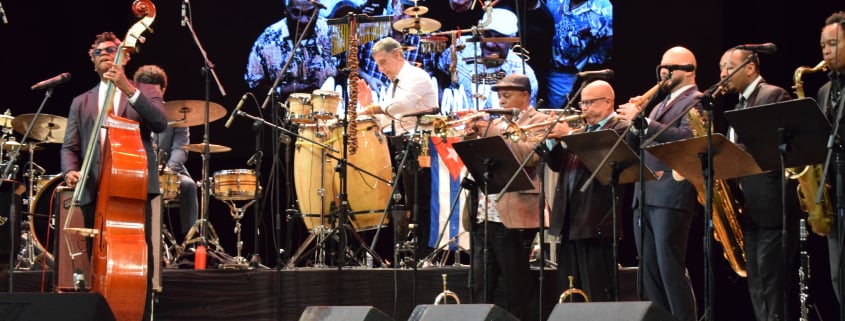 A large orchestra or big band performance on a stage, featuring musicians playing a double bass, drums, congas, trumpet, saxophone, and other brass instruments. A large projected image of smiling musicians is visible on the backdrop. A Cuban flag is visible behind the percussion.