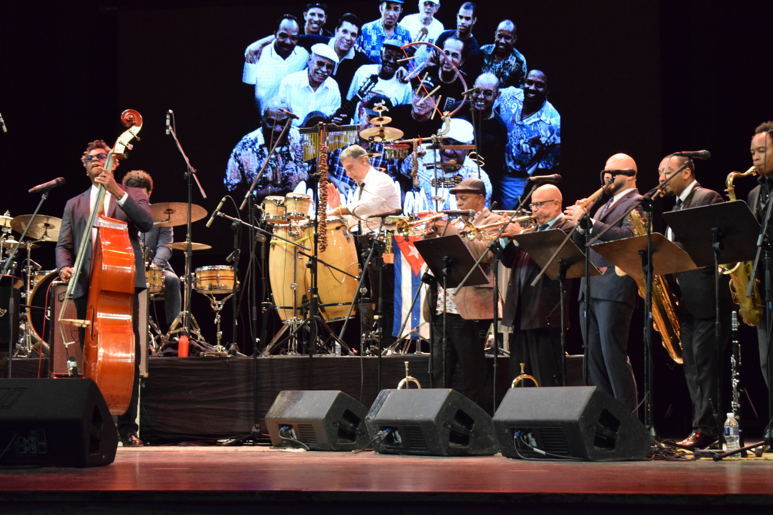 A large orchestra or big band performance on a stage, featuring musicians playing a double bass, drums, congas, trumpet, saxophone, and other brass instruments. A large projected image of smiling musicians is visible on the backdrop. A Cuban flag is visible behind the percussion.