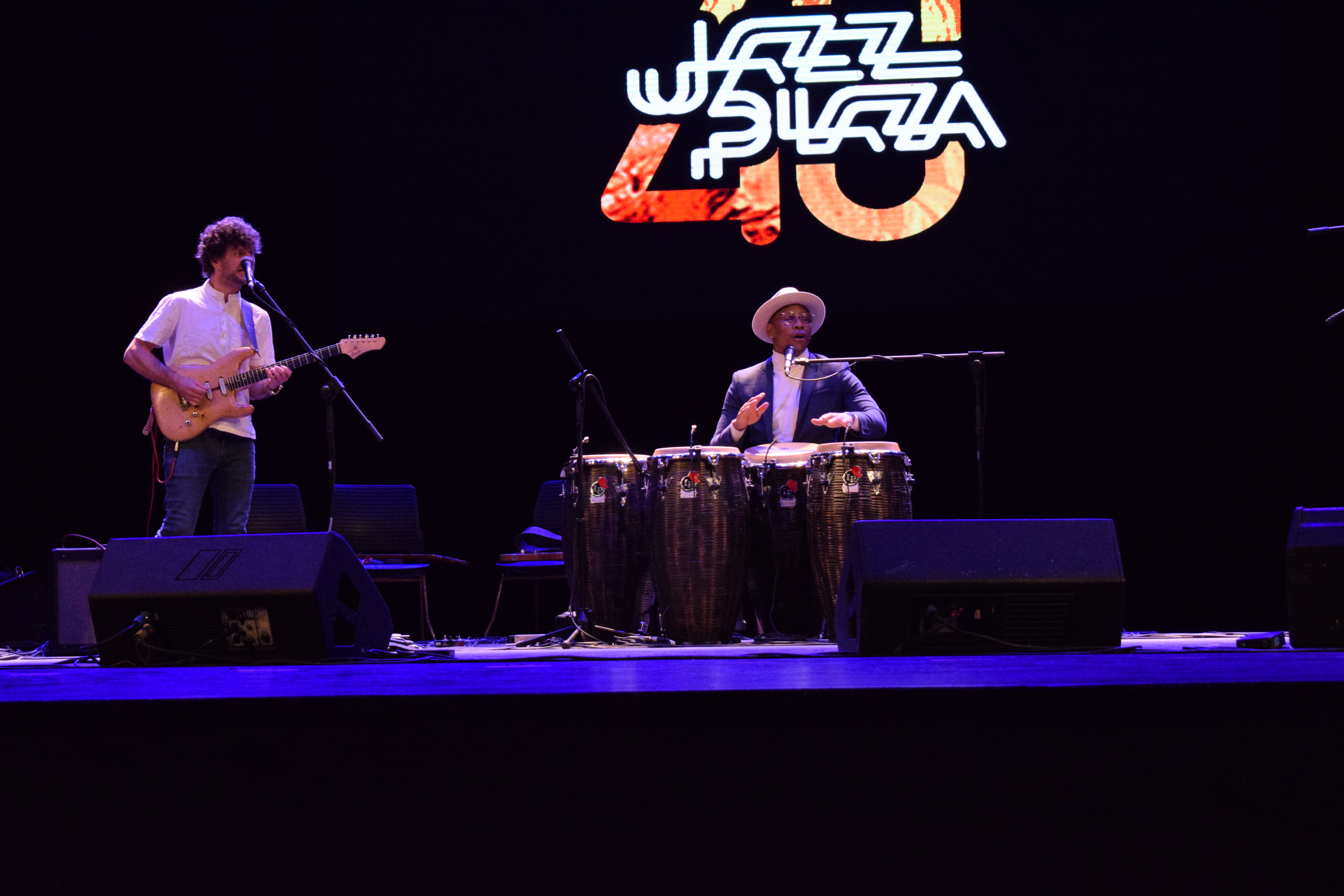 Two musicians on a stage under a logo reading "JAZZ PLAZA 40". One musician is standing and playing an electric guitar, and the other is seated behind a set of conga drums, wearing a suit and a fedora.
