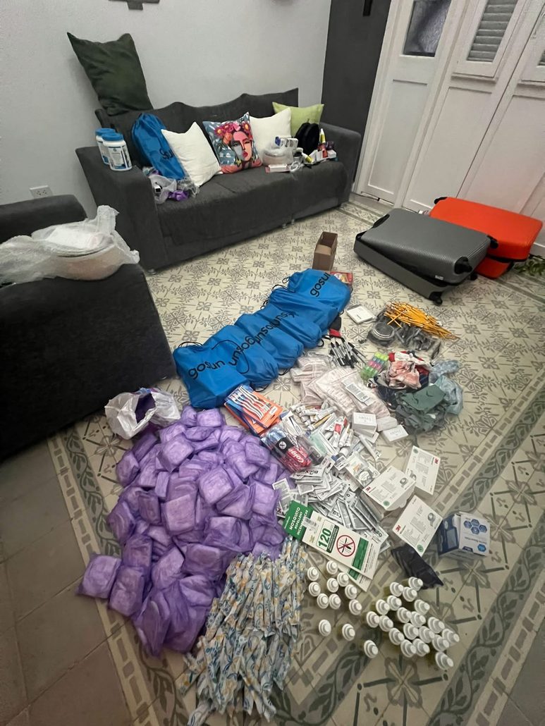 Bags, medical supplies and other donated items arranged on a tiled floor beside suitcases